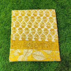 Peut inclure: Un tissu en coton blanc avec un imprim&eacute; floral jaune. Le tissu a une bordure avec un motif floral plus petit. Le tissu est pli&eacute; et pos&eacute; sur une surface herbeuse verte.