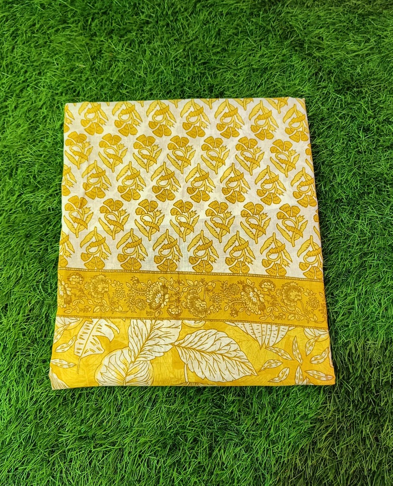 Peut inclure: Un tissu en coton blanc avec un imprim&eacute; floral jaune. Le tissu a une bordure avec un motif floral plus petit. Le tissu est pli&eacute; et pos&eacute; sur une surface herbeuse verte.