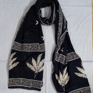 May include: A dark navy blue scarf with a white floral and leaf pattern. The scarf has a decorative border with a repeating design. The scarf is made of a lightweight fabric and is suitable for various occasions.