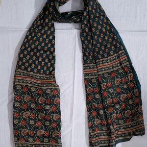 May include: Dark teal scarf with a floral pattern in red and beige. The scarf has a border with a repeating design. The scarf is made of a soft, flowing fabric, perfect for adding a touch of elegance to any outfit.