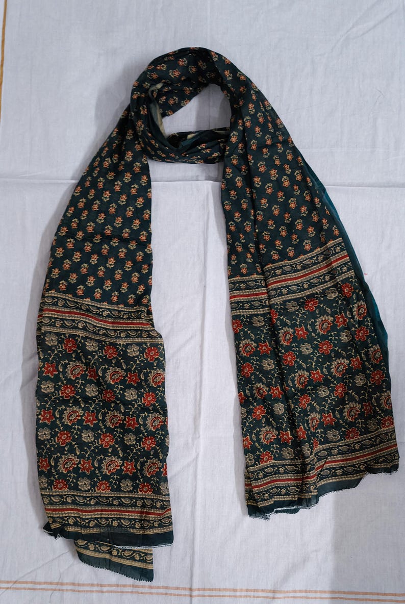 May include: Dark teal scarf with a floral pattern in red and beige. The scarf has a border with a repeating design. The scarf is made of a soft, flowing fabric, perfect for adding a touch of elegance to any outfit.