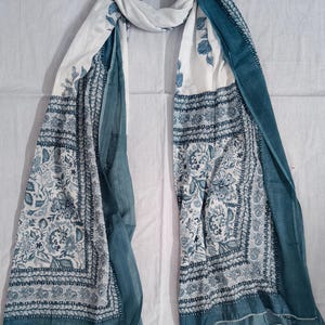 May include: A white and teal scarf with a floral pattern. The scarf has a rectangular shape with a border design and teal edges. The floral pattern is in shades of teal and blue. The scarf is draped on a white surface.