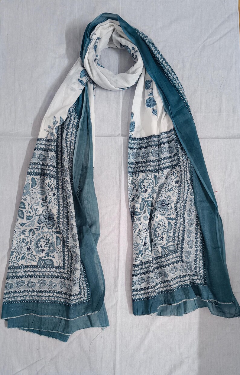 May include: A white and teal scarf with a floral pattern. The scarf has a rectangular shape with a border design and teal edges. The floral pattern is in shades of teal and blue. The scarf is draped on a white surface.
