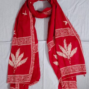 May include: A vibrant red scarf with intricate white floral and geometric patterns. The scarf features large, stylized leaf motifs and decorative borders. The fabric is draped, showcasing its flowing texture and design.