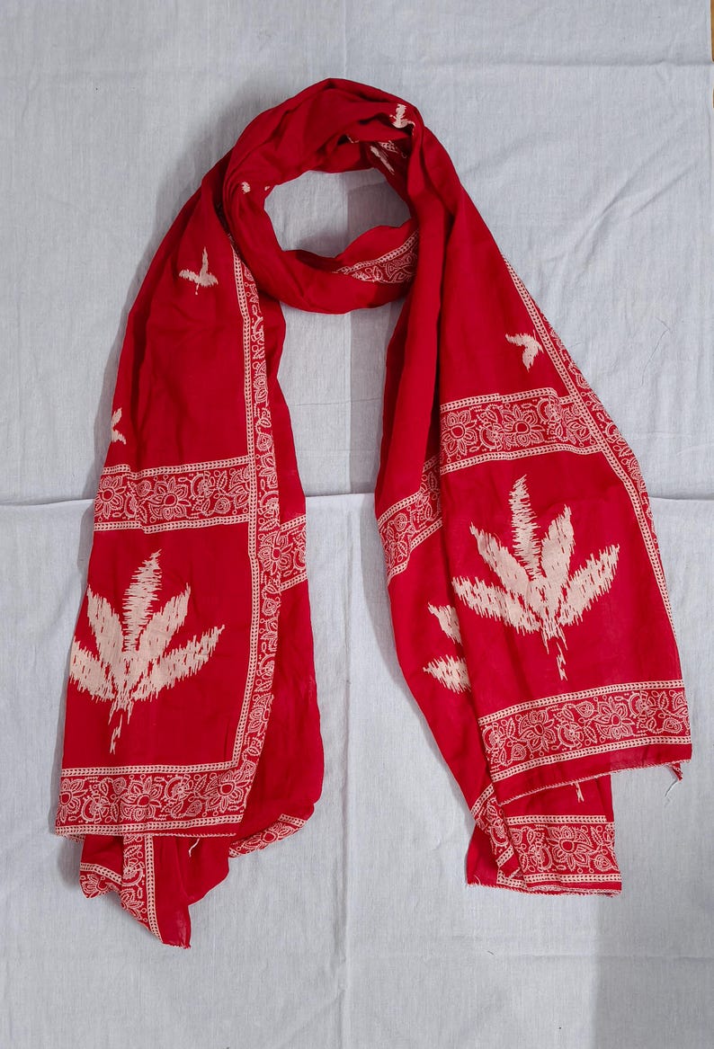 May include: A vibrant red scarf with intricate white floral and geometric patterns. The scarf features large, stylized leaf motifs and decorative borders. The fabric is draped, showcasing its flowing texture and design.