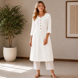 May include: A solid white knee-length tunic dress featuring three-quarter length sleeves, a notched neckline, and a vertical placket with four brown buttons. The garment is paired with matching white straight-leg trousers and brown open-toe sandals.