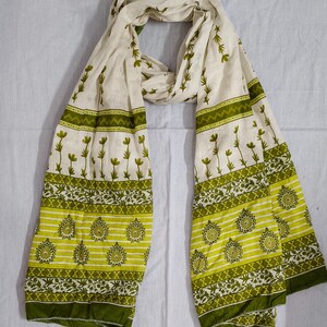May include: A cream-colored scarf with a repeating floral pattern in shades of green. The scarf has a border of green and yellow stripes with floral and geometric designs. The scarf is made of a lightweight fabric and is draped on a white surface.