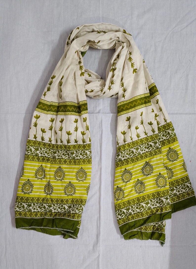 May include: A cream-colored scarf with a repeating floral pattern in shades of green. The scarf has a border of green and yellow stripes with floral and geometric designs. The scarf is made of a lightweight fabric and is draped on a white surface.