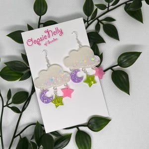May include: Cloud-shaped earrings with silver hooks, featuring white glitter clouds, purple crescent moons, pink stars, and green glitter stars. The earrings are on a white card with the text "Oopsie Nelly of Sweden".