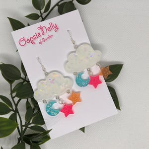 May include: A pair of white cloud-shaped earrings with iridescent glitter. Each earring has three charms: a blue crescent moon, a pink star, and an orange star.