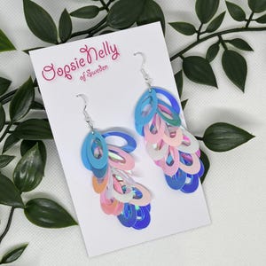 Può includere: Orecchini pendenti con paillettes ovali iridescenti stratificate nei toni del blu, rosa e bianco, fissati a ganci color argento. Gli orecchini sono esposti su un cartoncino bianco con il testo Oopsie Nelly of Sweden.