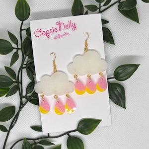 Può includere: Un paio di orecchini a forma di nuvola con ganci color oro. Ogni orecchino presenta una nuvola bianca e glitterata con tre ciondoli a goccia rosa e gialli. Gli orecchini sono esposti su una carta bianca con il testo "Oopsie Nelly of Sweden".