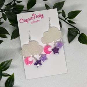 Può includere: Un paio di orecchini pendenti stravaganti con forme di nuvole bianche glitterate, lune crescenti rosa e stelle viola e viola scuro glitterate. Il testo "Oopsie Nelly of Sweden" è visibile.