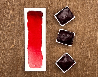 May include: Alizarine Crimson Dark watercolor paint in a pan. The paint is a deep red color and is shown in a white pan with a white label. The pan is sitting on a wooden surface.