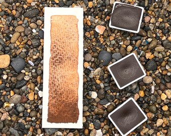 May include: A watercolour paint sample on a white paper card with three pans of brown watercolour paint on a bed of small brown and grey stones.