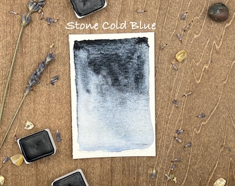 May include: A watercolor paint pan with the text "Stone Cold Blue" on a wooden surface with lavender sprigs and small stones.