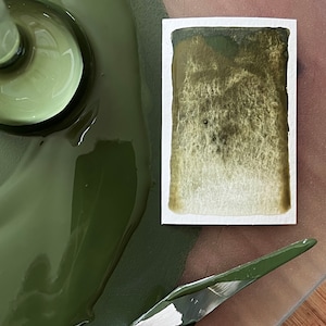 May include: A close-up of a green paint palette with a metal spatula and a small sample of the paint on a white card.