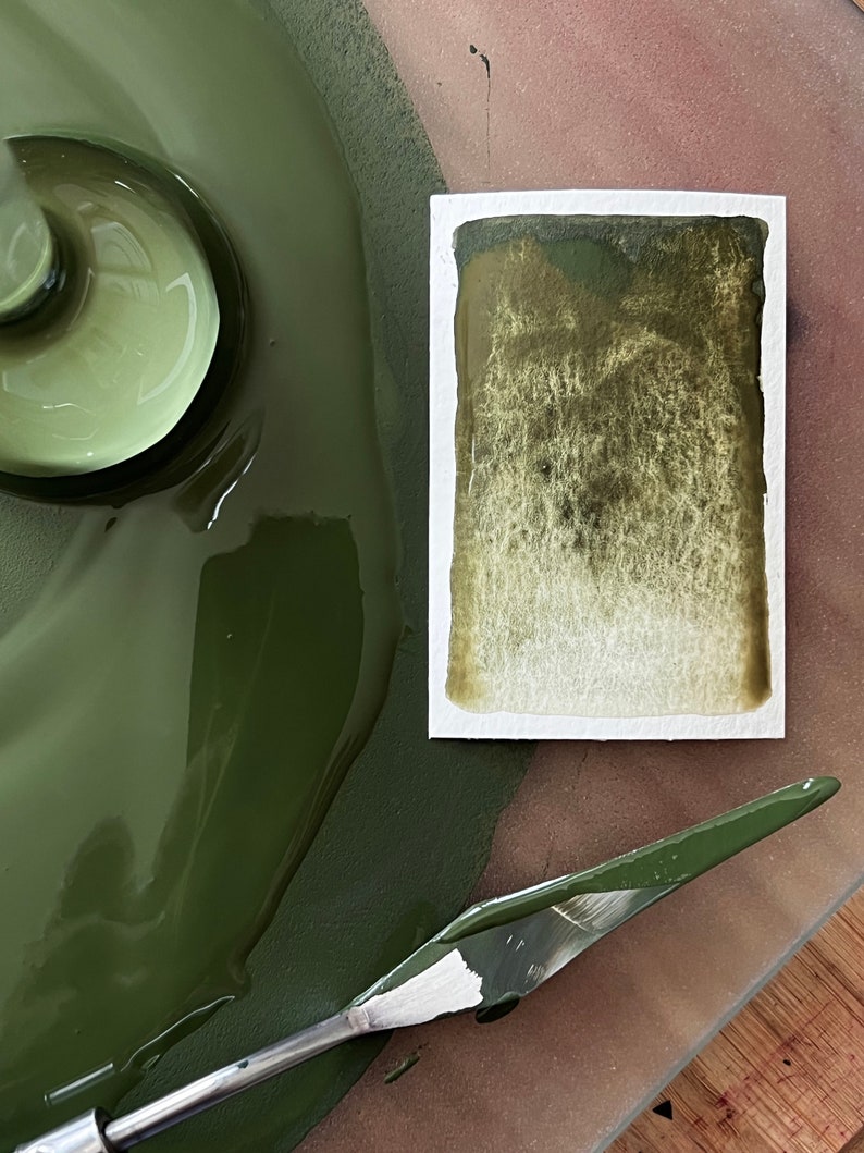 May include: A close-up of a green paint palette with a metal spatula and a small sample of the paint on a white card.