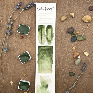 May include: A watercolor paint pan set with the name "Dusky Forest" written on the label. The pan is divided into three sections, each showing a different shade of green watercolor paint.