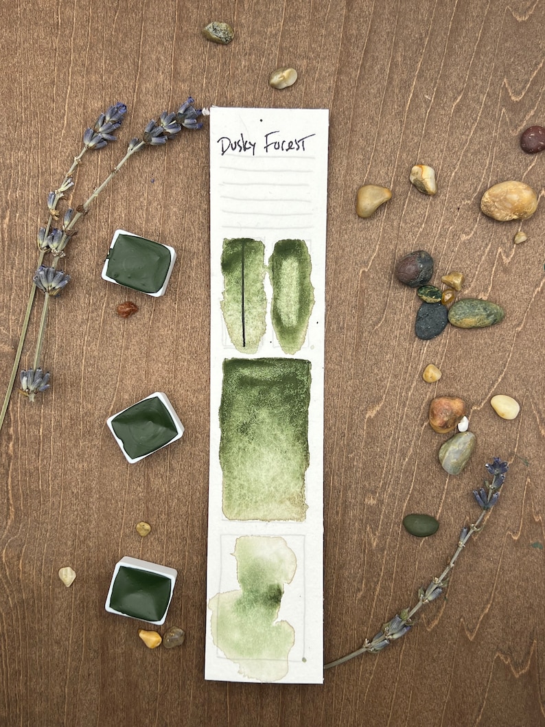 May include: A watercolor paint pan set with the name "Dusky Forest" written on the label. The pan is divided into three sections, each showing a different shade of green watercolor paint.