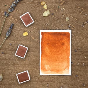 Könnte beinhalten: Vier Aquarellfarben in weißen Schalen mit brauner Farbe darin. Die Schalen befinden sich auf einer Holzoberfläche mit einem gemalten Aquarell-Farbverlauf in einem warmen Orangeton auf weißem Papier.