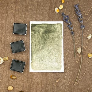 May include: Three black watercolor paint pans on a wooden surface with a watercolor wash on paper, dried lavender sprigs, and small yellow stones.