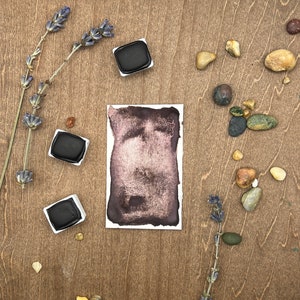 Könnte beinhalten: Drei schwarze Aquarellfarben auf einer Holzoberfläche mit getrockneten Lavendelzweigen und kleinen Steinen. Die Farbtöpfe sind in einem Halbkreis um ein Quadrat aus Aquarellpapier mit einem dunkelvioletten Aquarellwasch angeordnet.
