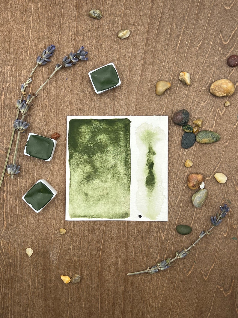 May include: A watercolor paint set with three pans of green paint. The pans are arranged in a circle on a wooden surface with lavender sprigs and small stones.