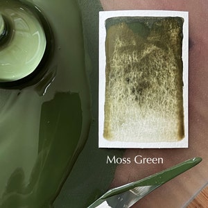 Op de afbeelding: Een close-up van een groen verfstaal met een wit label met de tekst "Moss Green". De verf is uitgesmeerd op een oppervlak met een witte spatel.