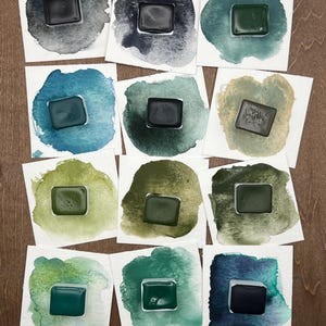 K&ouml;nnte beinhalten: Eine Sammlung von Aquarellfarbenmustern auf wei&szlig;en Karten. Jede Karte zeigt ein Quadrat aus dunkler, ges&auml;ttigter Farbe in verschiedenen Gr&uuml;n-, Blau- und Graut&ouml;nen, umgeben von einer Aquarellw&auml;sche. Die Farben werden auf einer Holzoberfl&auml;che pr&auml;sentiert.