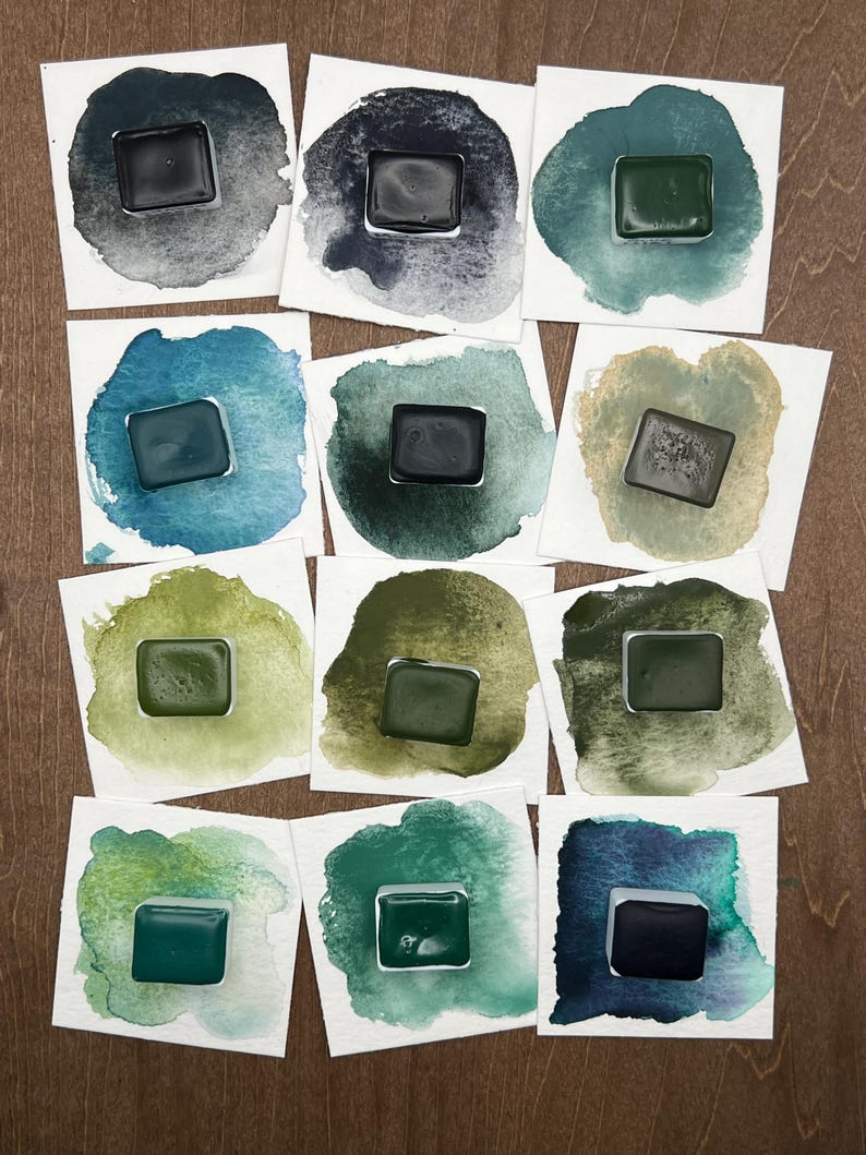 K&ouml;nnte beinhalten: Eine Sammlung von Aquarellfarbenmustern auf wei&szlig;en Karten. Jede Karte zeigt ein Quadrat aus dunkler, ges&auml;ttigter Farbe in verschiedenen Gr&uuml;n-, Blau- und Graut&ouml;nen, umgeben von einer Aquarellw&auml;sche. Die Farben werden auf einer Holzoberfl&auml;che pr&auml;sentiert.