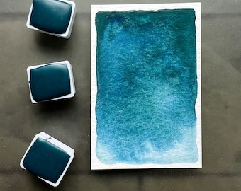 Può includere: Tre pastiglie di vernice ad acquerello in contenitori di plastica bianca con vernice blu teal scuro all'interno. Un campione di vernice è mostrato su una carta bianca con il testo "Peacock Blue".