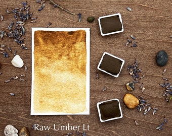 May include: A watercolour paint pan with a light brown colour swatch and the text "Raw Umber Lt" on a wooden surface with small stones and dried lavender flowers.