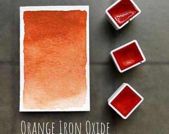 May include: A watercolor paint pan with a gradient of orange and red shades. The pan is labeled "ORANGE IRON OXIDE".