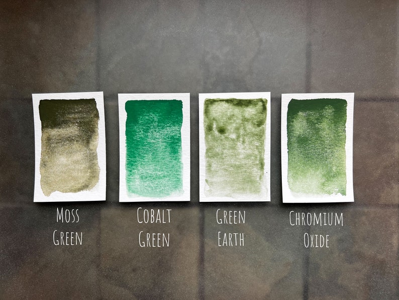 May include: Four watercolor paint swatches on white paper cards. The swatches are labeled "Moss Green", "Cobalt Green", "Green Earth", and "Chromium Oxide".