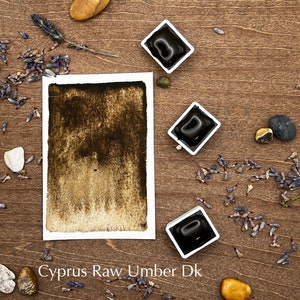 May include: A watercolor paint pan with a dark brown pigment labeled "Cyprus Raw Umber Dk". The pan is surrounded by dried lavender flowers and small stones on a wooden surface.