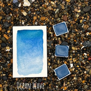 Op de afbeelding: Een aquarelverfpan met een blauwe gradi&euml;nt, met het label "Ocean Wave". De pan is omringd door kleine, donkere kiezelsteentjes.