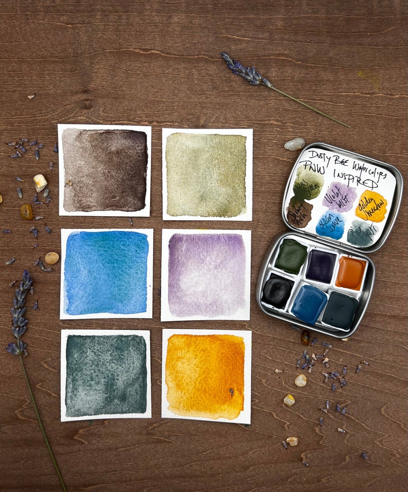 Op de afbeelding: Een set van zes aquarelverfstaaltjes in tinten bruin, groen, blauw, paars, grijs en geel. De staaltjes zijn gerangschikt in twee rijen van drie. De verf zit in een metalen blik met de woorden "Dusty Bee Watercolors PNW Inspired" op het deksel. Het blik is open en de verfpotjes zijn zichtbaar.