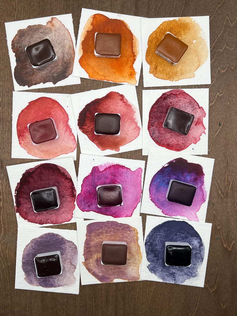 K&ouml;nnte beinhalten: Eine Sammlung von Aquarellfarbmustern auf quadratischem, wei&szlig;em Papier. Jedes Muster zeigt einen kleinen, dunkelbraunen Farbkasten in der Mitte, umgeben von einer lebendigen Farbwaschung. Die Farben reichen von warmen Orangen und Rot bis zu k&uuml;hlen Lila- und Blaut&ouml;nen.
