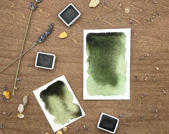 May include: Watercolor paint set with three dark green paint samples on white paper, and three square paint pans. The image also includes lavender sprigs, small stones, and dried lavender buds, all arranged on a wooden surface.