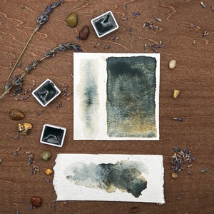 May include: Watercolor paint swatches in shades of dark blue and gray on textured paper. Two square paint pans and dried lavender sprigs are arranged on a wooden surface with small stones.