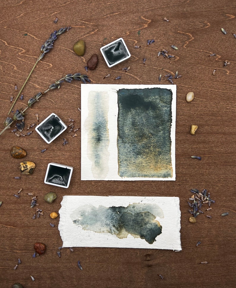 May include: Watercolor paint swatches in shades of dark blue and gray on textured paper. Two square paint pans and dried lavender sprigs are arranged on a wooden surface with small stones.
