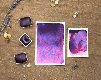 May include: Three watercolor paint pans in a purple hue, next to two watercolor paintings on white paper. The paintings are a gradient of purple and pink with a speckled texture. The background is a wooden surface with small stones and dried lavender.