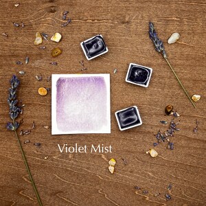 Op de afbeelding: Een aquarelverfpan met een paarse aquarelwas op een houten oppervlak met lavendeltakjes en kleine stenen. De tekst "Violet Mist" staat onder de verfpan.