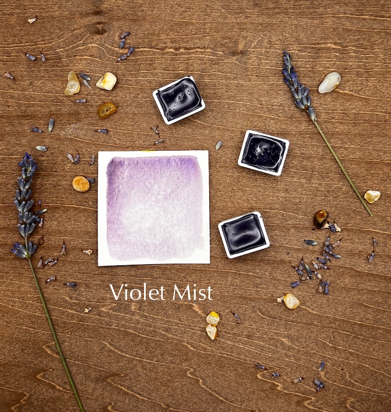 Op de afbeelding: Een aquarelverfpan met een paarse aquarelwas op een houten oppervlak met lavendeltakjes en kleine stenen. De tekst "Violet Mist" staat onder de verfpan.