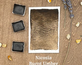 May include: Three watercolour paint pans in black, a sample of burnt umber watercolour paint, and the text "Nicosia Burnt Umber" on a wooden surface.