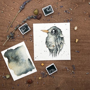 May include: Watercolor painting of a bird with large eyes and the text "Kiss Me". The artwork is on a white square paper, surrounded by watercolor paint, lavender, and small stones. The bird is gray with a yellow beak.