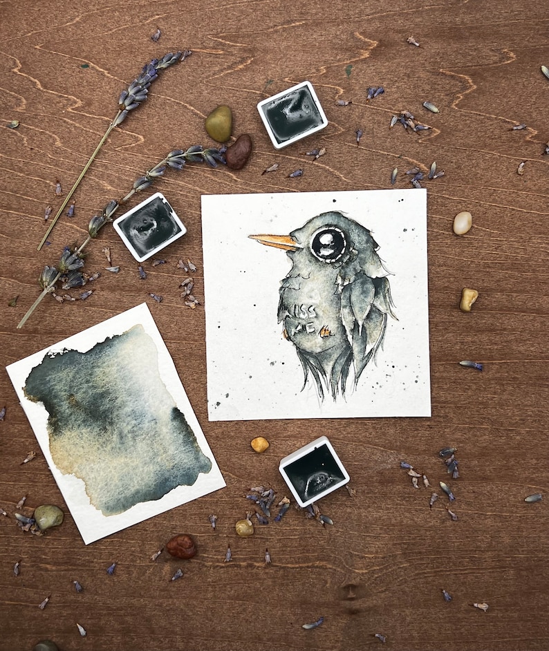 May include: Watercolor painting of a bird with large eyes and the text "Kiss Me". The artwork is on a white square paper, surrounded by watercolor paint, lavender, and small stones. The bird is gray with a yellow beak.
