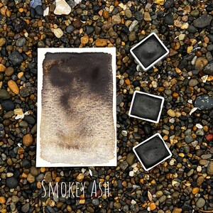 Op de afbeelding: Een aquarelverloop in rokerig asgrijs op een witte papieren achtergrond. Drie bakjes aquarelverf in zwarte plastic containers zijn rond het papier gerangschikt. De tekst "SMOKEY ASH" staat onder het papier.