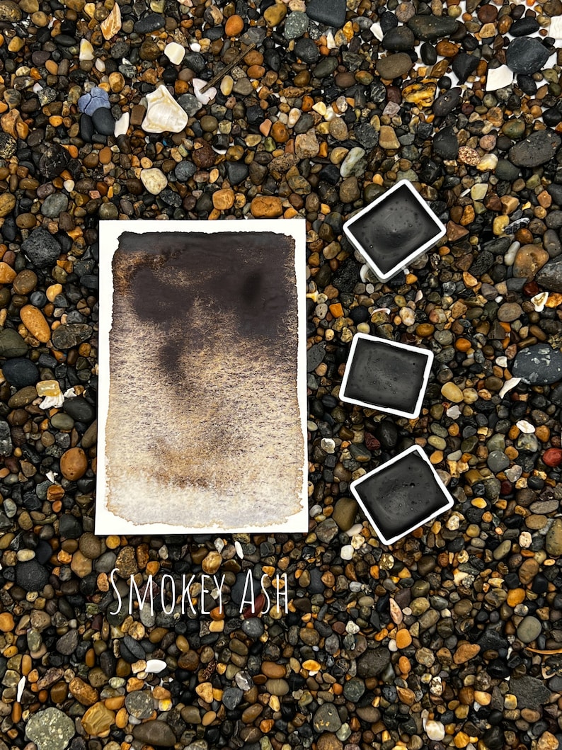 Op de afbeelding: Een aquarelverloop in rokerig asgrijs op een witte papieren achtergrond. Drie bakjes aquarelverf in zwarte plastic containers zijn rond het papier gerangschikt. De tekst "SMOKEY ASH" staat onder het papier.